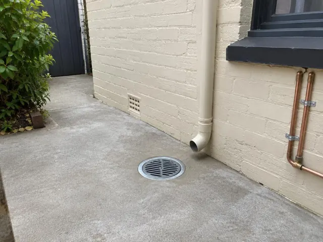 Outdoor grated drain beside a cream brick wall and concrete paving in Paddington